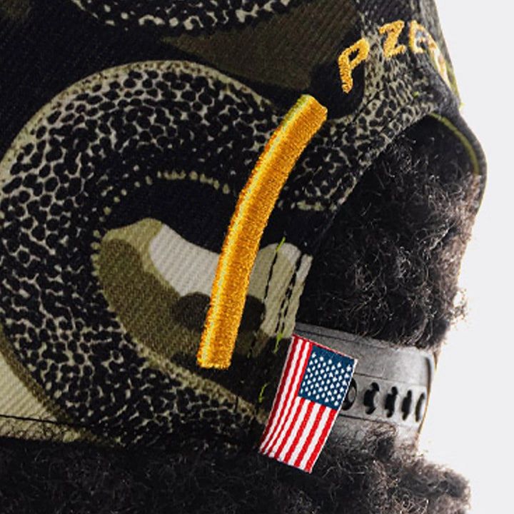 Close-up of a black and olive green camouflage 2026 Austin Formula 1 United States Grand Prix hat with a textured pattern, a gold embroidered strap, and a small American flag patch.