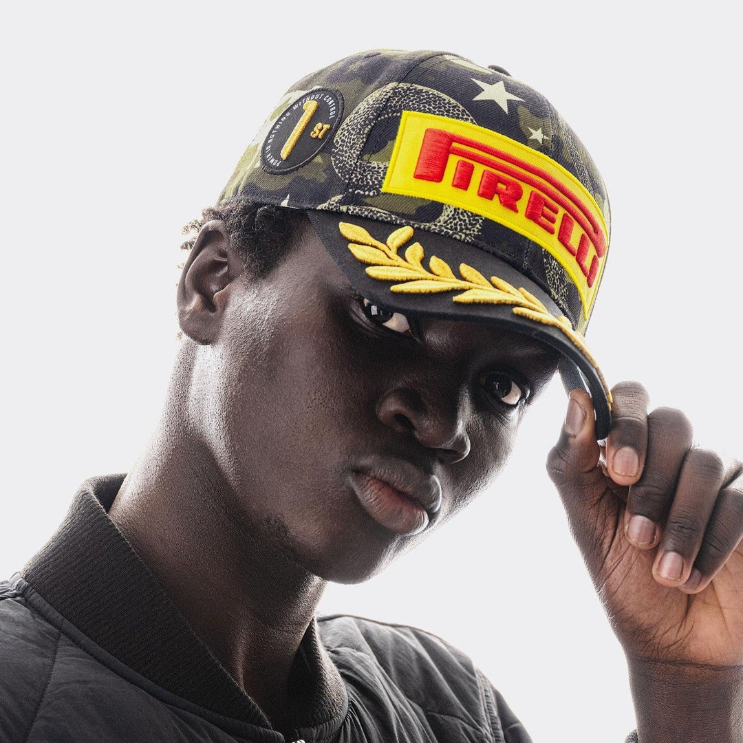 A close-up portrait of a man wearing a Official Pirelli Austin 2026 Podium Cap . The cap features a dark camouflage pattern with a large yellow and red Pirelli logo patch on the front, gold embroidered laurel leaves along the brim&
