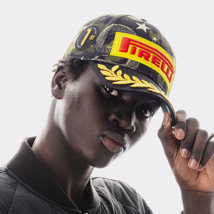 A close-up portrait of a man wearing a Official Pirelli Austin 2026 Podium Cap . The cap features a dark camouflage pattern with a large yellow and red Pirelli logo patch on the front, gold embroidered laurel leaves along the brim&
