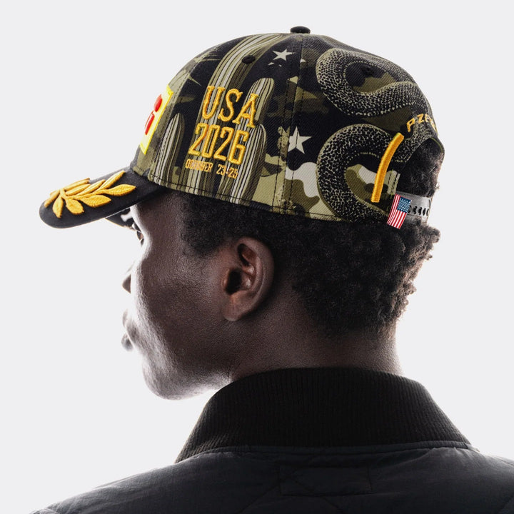 A model shown from the side wearing a black baseball cap with a camouflage pattern, a coiled rattle snake graphic, and yellow text reading &