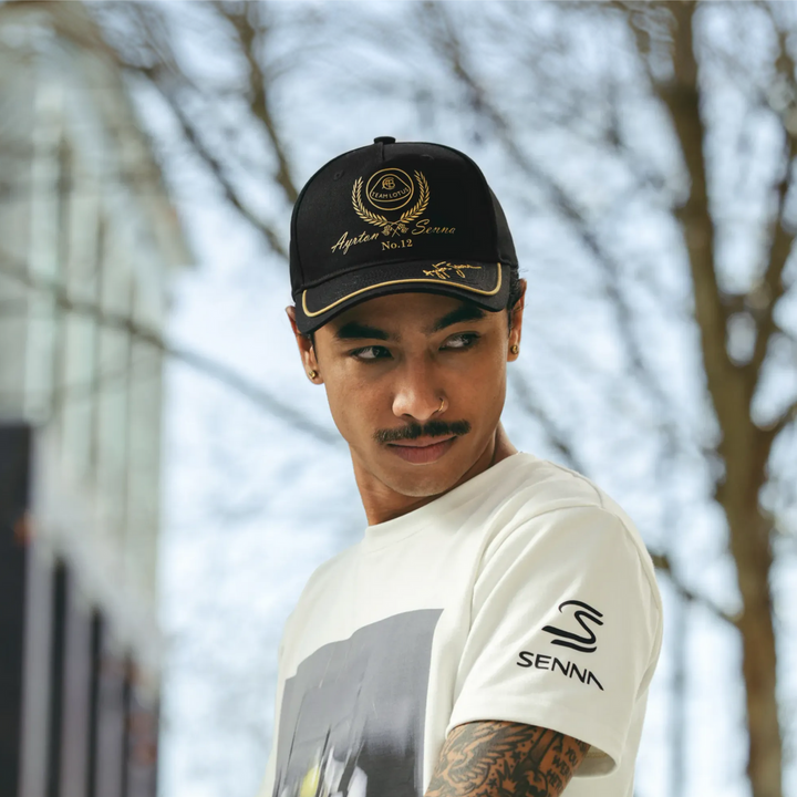 A man wearing a black and gold Senna Team Lotus F1 Racing Team cap and a white Senna t-shirt, photographed outdoors against a blurred background of trees and a building.