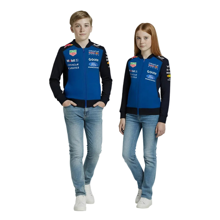 Two child models, a boy and a girl, wearing matching blue and black 2026 Red Bull F1 Team Racing full zip-up hooded sweatshirts with sponsor logos, paired with blue jeans and white sneakers, against a plain white background.